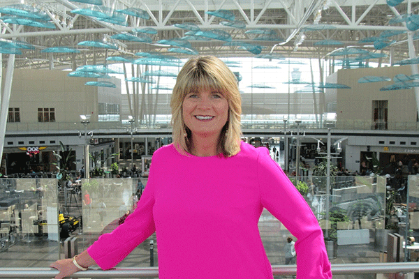 A woman with blonde hair and dressed in a pink blouse smiles directly at the camera. She is standing in an elevated position in front of an airport terminal.