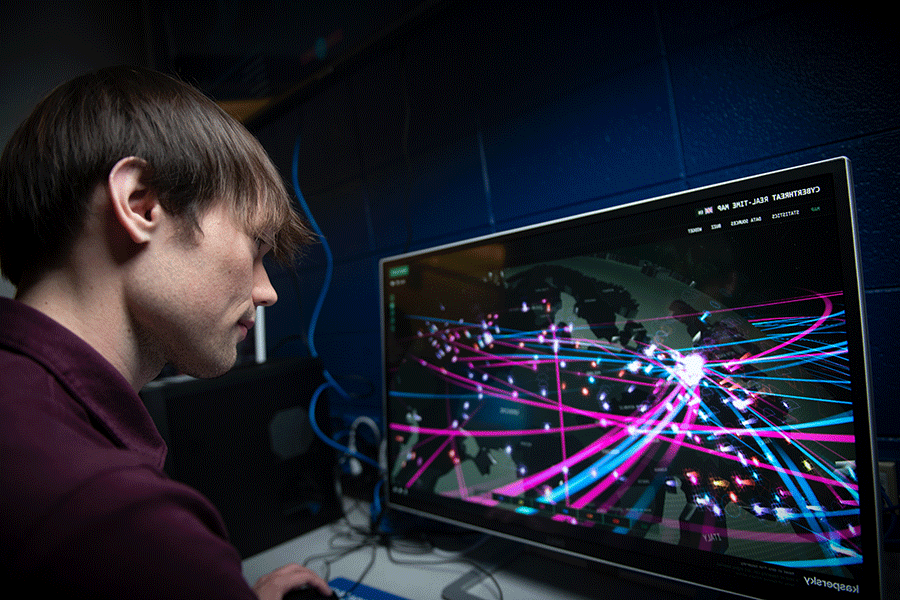 A white male student with short brown hair and wearing a black shirt is working on a computer. The monitor displays a colorful map of simulated cyberattacks. 