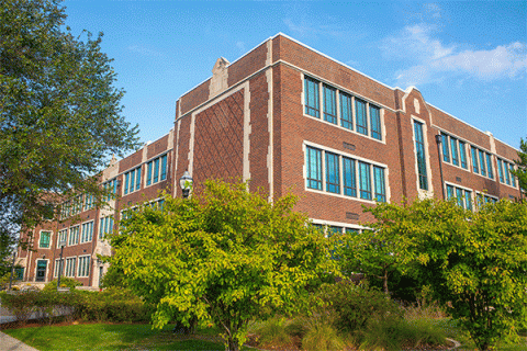 Exterior image of a large, multi-story brick building that houses the Bayh College of Education with green trees obscuring the lower half of the image with a blue sky.  